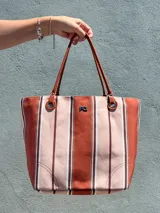 Cartera tipo tote bag confeccionada en canvas con estampado de rayas verticales en tonos beige, marrón y terracota. Cuenta con asas de hombro de cuero sintético marrón, detalles de ojalillos metálicos, cierre superior con broche magnético y logo metálico frontal.
