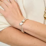 Pulsera de aleación de metales bañados en plata, compuesta por dos tiras de cuentas metálicas y detalles de cuero marrón oscuro, con un adorno central de dos gotas plateadas.