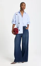 Camisa de algodón a rayas celestes y blancas, con cuello clásico, cierre frontal de botones, mangas largas y aberturas troqueladas en forma de corazón junto a bordados decorativos y texto en el hombro.