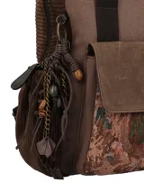 Mochila marrón de diseño patchwork y estilo bohemio, confeccionada en material textil. Cuenta con compartimento principal con cierre, bolsillo frontal con solapa decorada con tapicería estampada, bolsillos laterales, asas superiores fijas y correas ajustables. Incluye un llavero colgante lateral con borla y cuentas decorativas.