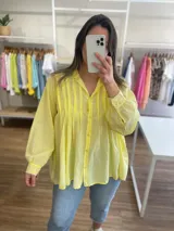 Camisa de algodón color amarillo con diseño de línea italiana, corte holgado y asimétrico. Presenta cuello camisero, cierre frontal con botones y detalle de pliegues verticales en el pecho. Cuenta con mangas largas y puños con botón.