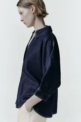Camisa de denim azul oscuro con corte amplio, cuello solapa, escote en pico y mangas tres cuartos. Presenta un aplique metálico dorado en el centro y bajo asimétrico.