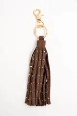 Charm para bolso compuesto por flecos de cuero sintético color marrón, decorados con pequeñas tachas metálicas doradas. Incluye un aro y un cierre de mosquetón metálico en tono dorado.