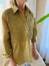 Camisa de manga larga color verde militar, con cuello clásico y cierre frontal de botones. Presenta un canesú bordado con diseño ornamental en la parte frontal superior.