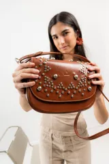 Cartera bandolera de cuero 100% natural color marrón, con estructura armada y tapa decorada con tachas plateadas colocadas a mano. Forrada en tela con bolsillo interno con cierre.