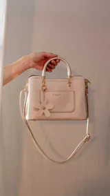 Cartera tipo tote color bordó, con doble asa corta y correa larga desmontable. Tiene bolsillo externo con solapa y dije colgante en forma de flor.