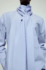 Camisa de algodón color celeste, con cuello alto y lazo integrado en el mismo tejido. Presenta cierre frontal con botones, manga larga con puños y un bolsillo frontal en el pecho. Diseño de corte recto.