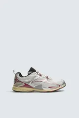 Zapatilla deportiva tipo running con diseño técnico, confeccionada en una combinación de materiales sintéticos y malla. Presenta una estética metalizada en tonos plata con detalles en color granate y una suela con volumen de diseño irregular en tono crema.
