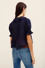 Camisa azul marino de algodón, con mangas cortas abullonadas y puños fruncidos con elástico. Presenta un amplio escote en V adornado con volados con detalles bordados y ojales a lo largo de la botonera frontal.