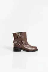 Botas de caña baja en cuero sintético color marrón, con diseño de estilo biker que incluye dos correas horizontales con hebillas metálicas y una argolla decorativa en el lateral. Posee suela y taco bajo de color negro.