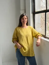 Blusa mostaza de lino, con cuello redondo, mangas tres cuartos con presilla y botón, y detalle de pliegues en la parte inferior.