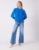 Blusa azul de gasa con pliegues en la parte delantera y detalles de encaje calado. Cuello alto fruncido y mangas largas con puños elastizados. Espalda lisa con abertura tipo gota y botón.