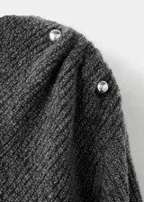 Jersey de punto gris con cuello barco y diseño de bajo asimétrico. Presenta manga larga y un detalle metálico decorativo en uno de los hombros.