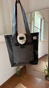 Bolso tote de ecocuero marrón oscuro con textura cocodrilo y un parche frontal de cuero liso marrón claro. Posee asas de hombro de cuero marrón y un broche decorativo blanco en el frente.