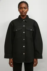 Sobrecamisa de jean color negro, con corte holgado, cuello clásico, cierre frontal con botones metálicos y dos bolsillos frontales con solapa.