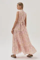 Vestido largo de algodón color rosa con estampado floral en tonos beige y dorado. Tiene cuello alto con lazo ajustable, corte imperio, falda escalonada con volados y diseño sin mangas.