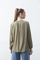 Camisa de color celeste claro con silueta relajada y mangas largas. Presenta un cuello redondo con pequeña abertura frontal y botones, además de un delicado trabajo de tablas en los hombros que aporta textura al diseño.