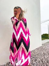 Vestido camisero largo con estampado de zig zag en colores fucsia, negro, blanco y rosa pálido. Tiene cuello camisero, mangas largas con puños abotonados y cierre frontal con botones.
