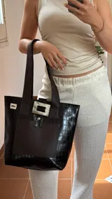 Bolso tote de ecocuero marrón oscuro con textura cocodrilo y un parche frontal de cuero liso marrón claro. Posee asas de hombro de cuero marrón y un broche decorativo blanco en el frente.