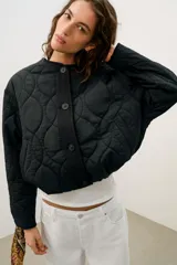Cazadora corta acolchada de color negro, con cuello redondo, manga larga y cierre frontal con botones. Presenta un diseño de costuras geométricas y bolsillos laterales de vivo.