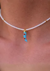 Collar tipo gargantilla compuesto por pequeñas cuentas de acrílico blancas, con dije de cuerno italiano (cornicello) esmaltado en turquesa y adornado con dos piedras rojas redondas. Los herrajes son dorados.