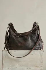 Cartera hobo de cuero color marrón oscuro con acabado gastado. Presenta una solapa frontal con detalles de ojales metálicos, una argolla metálica en la correa para el hombro y bolsillos laterales. Incluye un llavero colgante con candado y dijes.