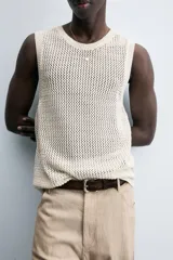 Chaleco de punto color blanco roto, con cuello redondo y sisa, confeccionado en tejido calado de estructura abierta y acabado en rib.