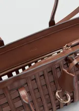 Bolso tipo cesta de color marrón, con diseño trenzado y estructura cuadriculada, asas cortas de mano y correa larga ajustable y desmontable. Incluye charm decorativo y cierre de cremallera.