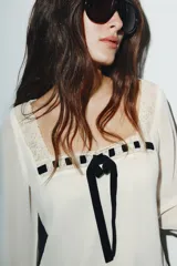 Camisa de georgette color blanco con escote cuadrado, mangas largas acampanadas y forro interior. Presenta detalles de puntilla a tono en el escote y un lazo de terciopelo negro en el centro del pecho.