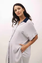 Vestido camisero maternal de color gris claro, con cuello tipo polo en contraste azul marino, cierre frontal con botones y bordado de un corazón pequeño en el pecho. Presenta mangas cortas y bolsillos laterales.