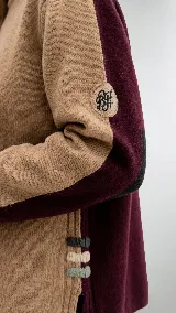 Polera de cuello alto confeccionada en lana lambswool, con diseño bicolor en negro y marrón. Presenta mangas largas con franjas verticales en gris y un parche circular bordado en la manga derecha.