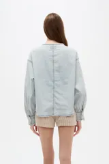 Blusa de jean ligera en color celeste claro, con cuello redondo y cierre frontal parcial con botones. Presenta un diseño con volados a lo largo de la botonera, bolsillo ojal en el pecho, y mangas largas abullonadas con puños ajustados y fruncidos.