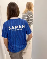 Camiseta de fútbol de manga corta en color azul con diseño estampado abstracto y cuello en V rosado con detalles blancos a juego con los puños. Presenta inscripciones y escudos deportivos en el pecho.