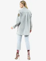 Chaqueta de denim en tono azul claro con efecto lavado, corte holgado y cierre frontal con botones. Presenta múltiples insignias decorativas, incluyendo broches con cristales, una insignia con cinta y detalles de jacquard en los laterales. Cuenta con bolsillos frontales y aberturas laterales.
