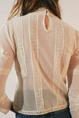 Blusa de manga larga color crema, con cuello alto y detalles de encaje y bordados florales. Presenta paneles verticales con entredoses y un tejido semitransparente.