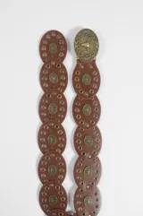 Cinturón de cuero marrón compuesto por piezas circulares unidas, decoradas con tachas metálicas doradas y apliques centrales con relieve de cabeza de toro. Incluye una hebilla metálica circular grande con diseño grabado.
