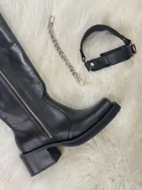 Bota alta tipo biker de cuero negro con cierre metálico lateral que recorre toda la caña. Presenta un diseño versátil con estribo y cadena metálica desmontables en la zona del tobillo, taco cuadrado y plataforma delantera.