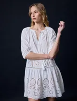 Vestido corto blanco de algodón con bordado calado tipo broderie, cuello redondo, escote en V con lazo y borlas, y mangas tres cuartos con puño.