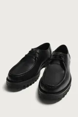 Zapato de cordones de estilo derby, color negro, con parte superior lisa, detalle de bordón en el empeine, horma redonda y suela track dentada.
