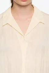 Camisa corta de corte boxy en color marrón, con cuello camisero, cierre frontal de botones y mangas 3/4. Presenta pespuntes decorativos a contraste en los puños y una textura sutil en el tejido.
