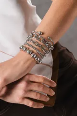 Conjunto de cinco pulseras plateadas de diseño variado que incluye una esclava abierta con terminaciones esféricas, eslabones de cadena gruesa y fina, una cadena de cuentas cuadradas y una pulsera de esferas metálicas.