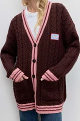 Cardigan de punto con diseño de bloques de color, combinando azul marino en el cuerpo y beige en las mangas. Presenta un tejido con textura de trenzas, cuello en V con borde rayado en azul y blanco, cierre frontal con botones y bolsillos delanteros. Incluye un parche decorativo en el pecho.