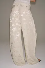 Pantalón de tiro medio confeccionado en hilatura con lino, con cintura con trabillas, bolsillos laterales ocultos y pierna ancha. Presenta un diseño con estampado jacquard de motivos florales en tonos beige y crudo, y cierre frontal con cremallera, botón y ganchos metálicos.
