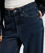 Pantalón de jean corte baggy con cintura media, diseño de cinco bolsillos y cierre frontal con cremallera y botón. Presenta un lavado oscuro sobretinta y una trabilla con colgante metálico desmontable.