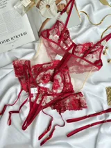 Set de lencería de tul transparente nude con bordado floral rojo, compuesto por un corset con sostén de aro y portaligas integrado, una tanga con apertura trasera y guantes cortos translúcidos con volados.