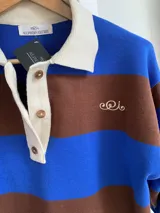 Buzo de tejido de punto con diseño a rayas horizontales anchas en color azul y marrón. Presenta cuello tipo polo en color crema con cierre frontal de botones y un pequeño bordado decorativo en el pecho.