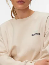 Buzo boxy marrón con cuello redondo, manga larga y diseño holgado, con pequeño estampado circular en el pecho.