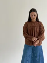 Blusa blanca de corte holgado con cuello redondo, mangas largas con puños elásticos y detalles de puntillas verticales en el frente.