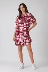 Vestido camisero de corte holgado y silueta relajada, con cuello clásico, cierre frontal de botones y mangas cortas con dobladillo. Presenta un estampado de cuadros irregulares en color bordó sobre fondo blanco.
