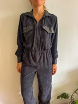 Mono largo de pana color negro, con cuello camisero y cierre frontal con cremallera. Presenta bolsillos con solapa de cuero en el pecho y puños de cuero.